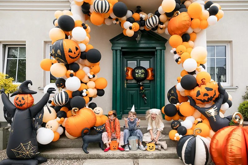 A Halloween decoration setup in front of a house door, featuring a large balloon garland arch in orange, black, and white, pumpkin decorations, a black witch figure, and three children sitting on the doorstep wearing costumes.