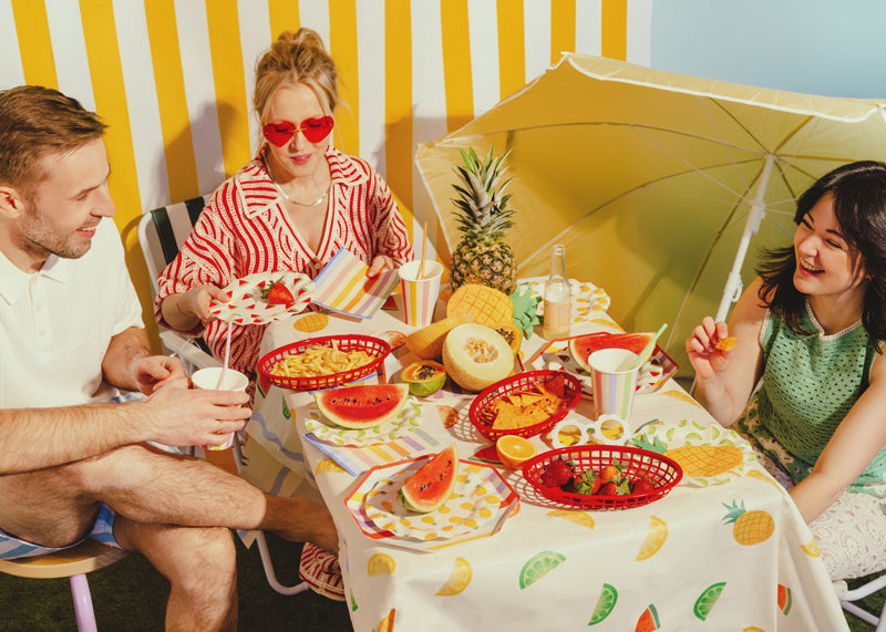 Three people sitting at a table with a colorful tablecloth and various fruits and drinks.