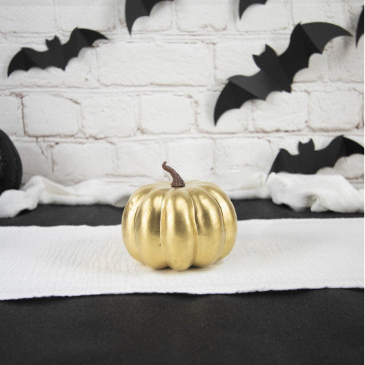 A shiny gold decorative pumpkin sits on a white runner in front of a white brick wall decorated with black bat cutouts.