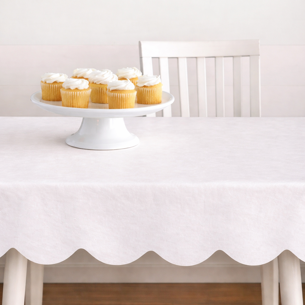 White scalloped tablecloth on a dining table with a white cake stand holding cupcakes.