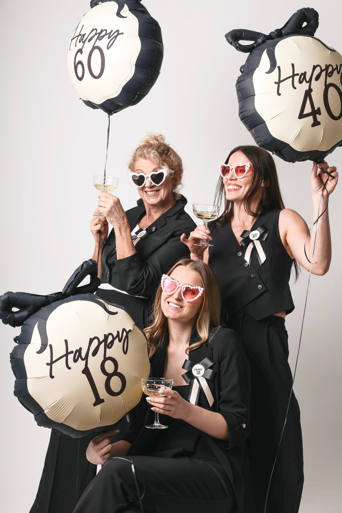 Three women holding 'Happy milestone' balloons with numbers, posing together against a plain background.