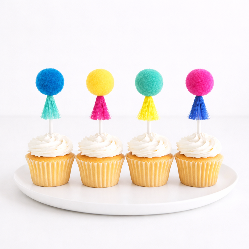 Four cupcakes with colorful pom-pom toppers on a white plate against a white background