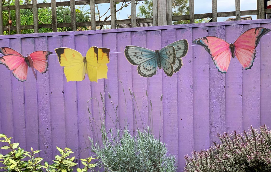 Butterfly-shaped decorations on a purple wooden fence with plants in the foreground.