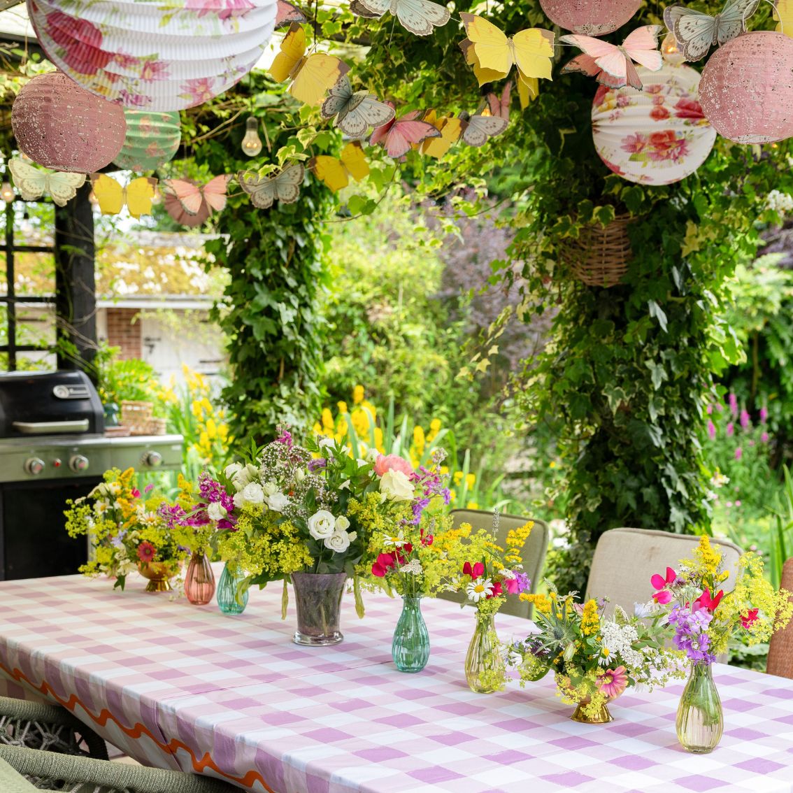 Outdoor garden party setup with floral arch, lanterns, and checkered tablecloth.