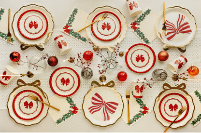 A festive christmas table setting featuring  plates with red bow motifs, gold-rimmed edges, and red and gold tableware, arranged on a white tablecloth with Christmas ornaments and greenery.
