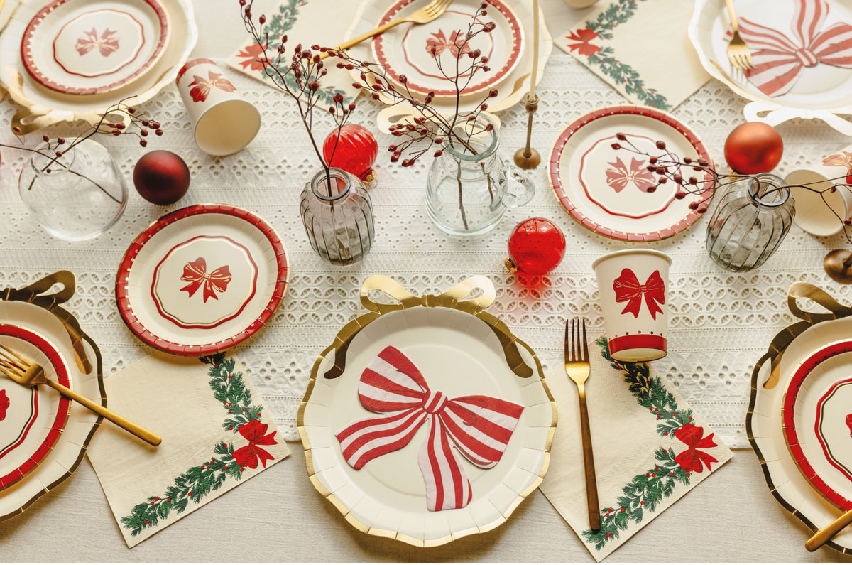 A festive tabletop setting with Christmas-themed paper plates, matching cups, gold-toned forks, lace tablecloth, red ornaments, and small glass vases with berry stems.
