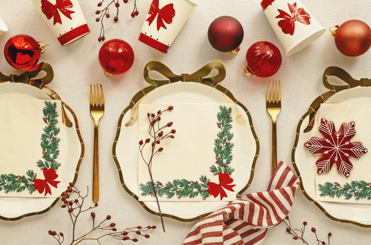 A festive holiday table setting featuring three round white plates with gold trim, decorated with green wreaths, red bows, and red berries. Each plate is paired with a gold fork and a matching napkin.
