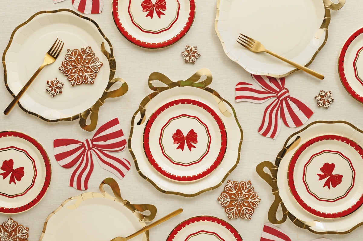 A festive holiday table setting featuring white plates with red and gold patterns, gold forks, red and white striped napkins, and gold snowflake decorations on a cream background.
