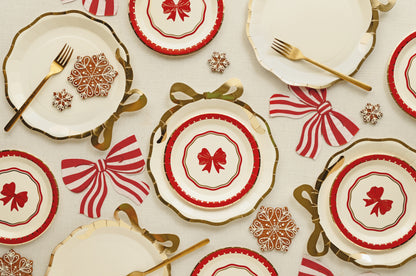 A festive holiday table setting featuring white plates with red and gold patterns, gold forks, red and white striped napkins, and gold snowflake decorations on a cream background.