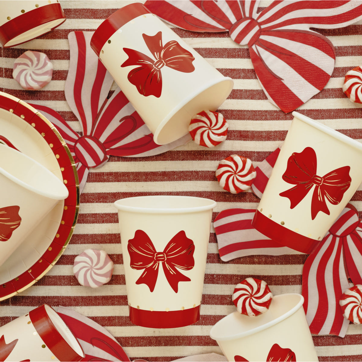Red and white holiday party tableware set featuring paper cups, plates, napkins, and bows with a red bow graphic on a red and white striped background.