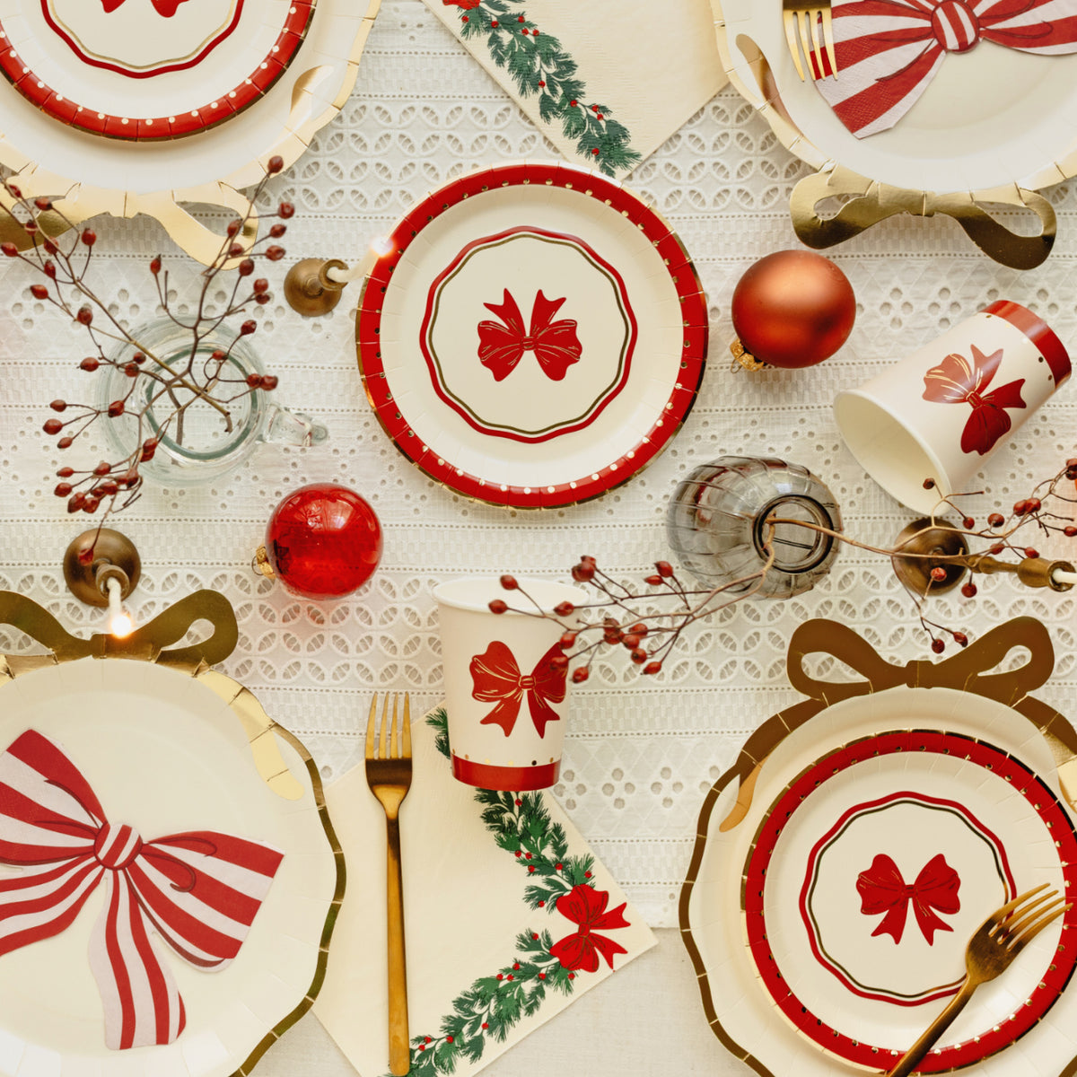 A festive holiday table setting featuring white paper plates and cups decorated with red bows, red-and-white striped accents, green holly leaves, gold forks, and red ornaments on a white lace tablecloth.