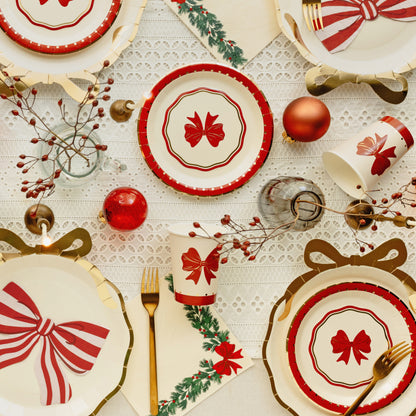 A festive holiday table setting featuring white paper plates and cups decorated with red bows, red-and-white striped accents, green holly leaves, gold forks, and red ornaments on a white lace tablecloth.
