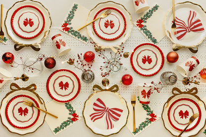 A festive holiday table setting featuring multiple white plates with red bow and floral motifs, gold-rimmed edges, matching cups, gold forks, green holly sprigs, red ornaments, and delicate dried floral accents arranged on a white textured tablecloth.