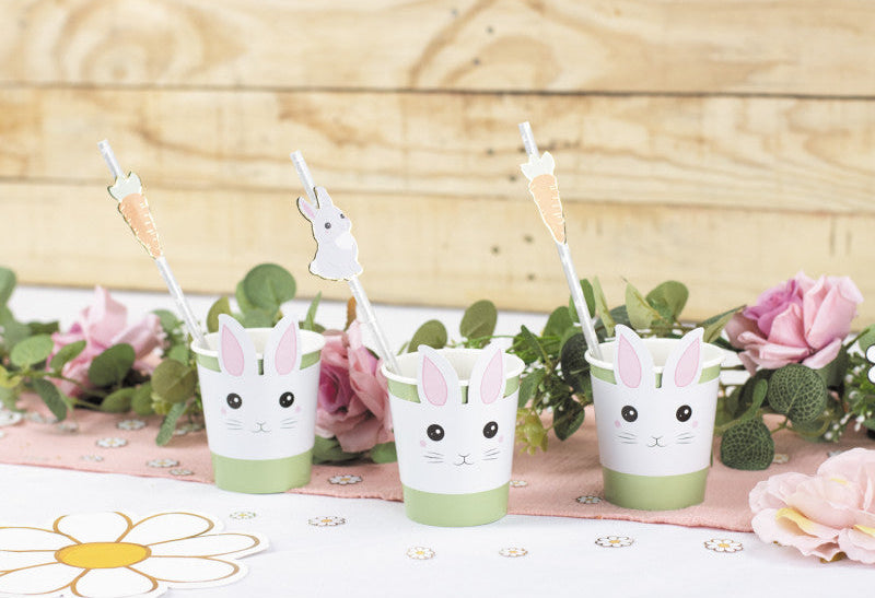 Three bunny-themed cups with straws on a tablecloth with flowers and a wooden background