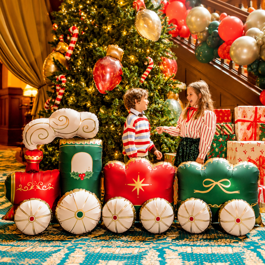 Giant red and green Christmas train decoration beside a decorated Christmas tree with presents and festive balloons.
