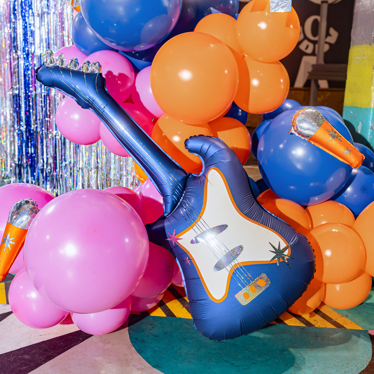 A blue inflatable electric guitar balloon decoration displayed among pink and orange balloons, with a silver tinsel backdrop and party decor in the background.
