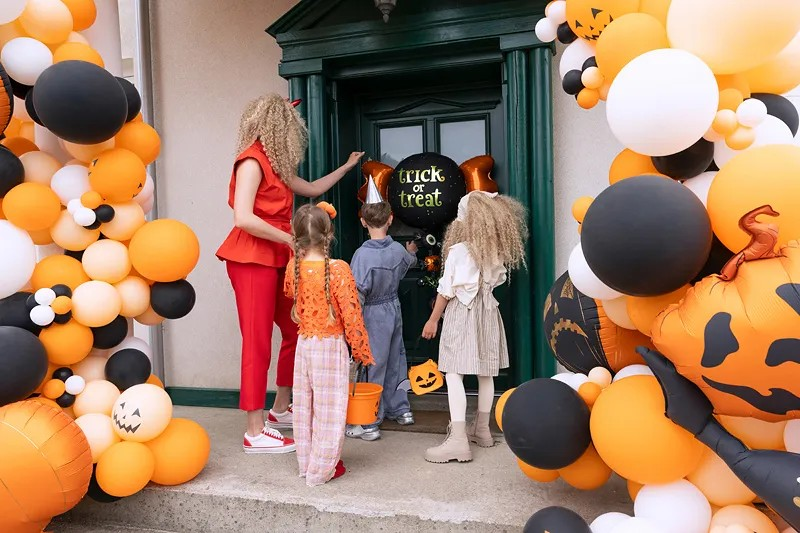 A family of four celebrates Halloween outside a house decorated with a large balloon arch of orange, black, and white balloons, pumpkins, and a 'Trick or Treat' sign hanging on the green front door.