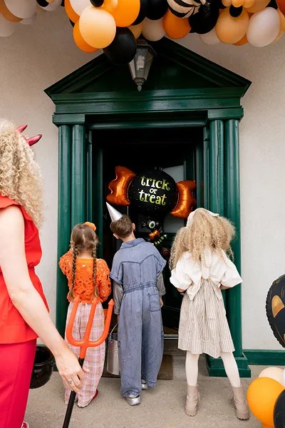 Three children in Halloween costumes stand at the doorway of a decorated house, looking inside toward a 'Trick or Treat' sign. The house has a dark green door frame, orange, black, and white balloons, and a pumpkin decoration.