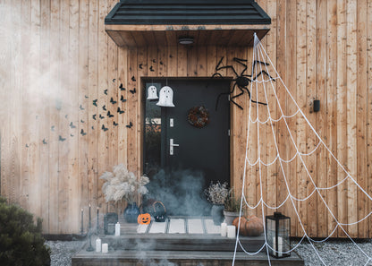 Halloween front porch decoration with a black door, wood siding wall, white spiderwebs, ghost decorations, black bats, a pumpkin, candles, and smoke effects.