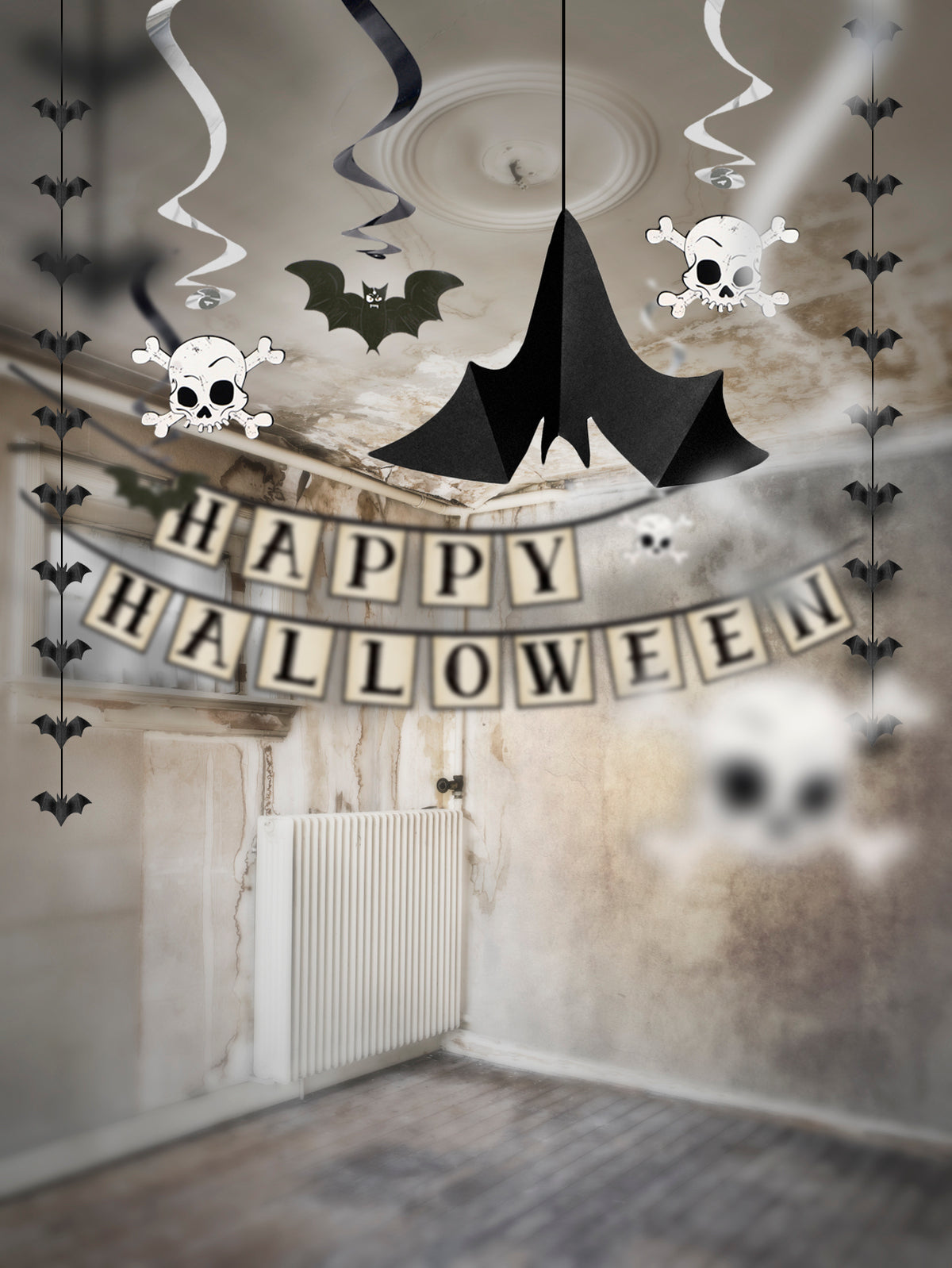 Halloween party decoration kit shown hanging in an empty room, featuring a black bat-shaped ceiling ornament, skull and crossbones wall decals, black bat ribbons, a 'HAPPY HALLOWEEN' banner, and white skull streamers.