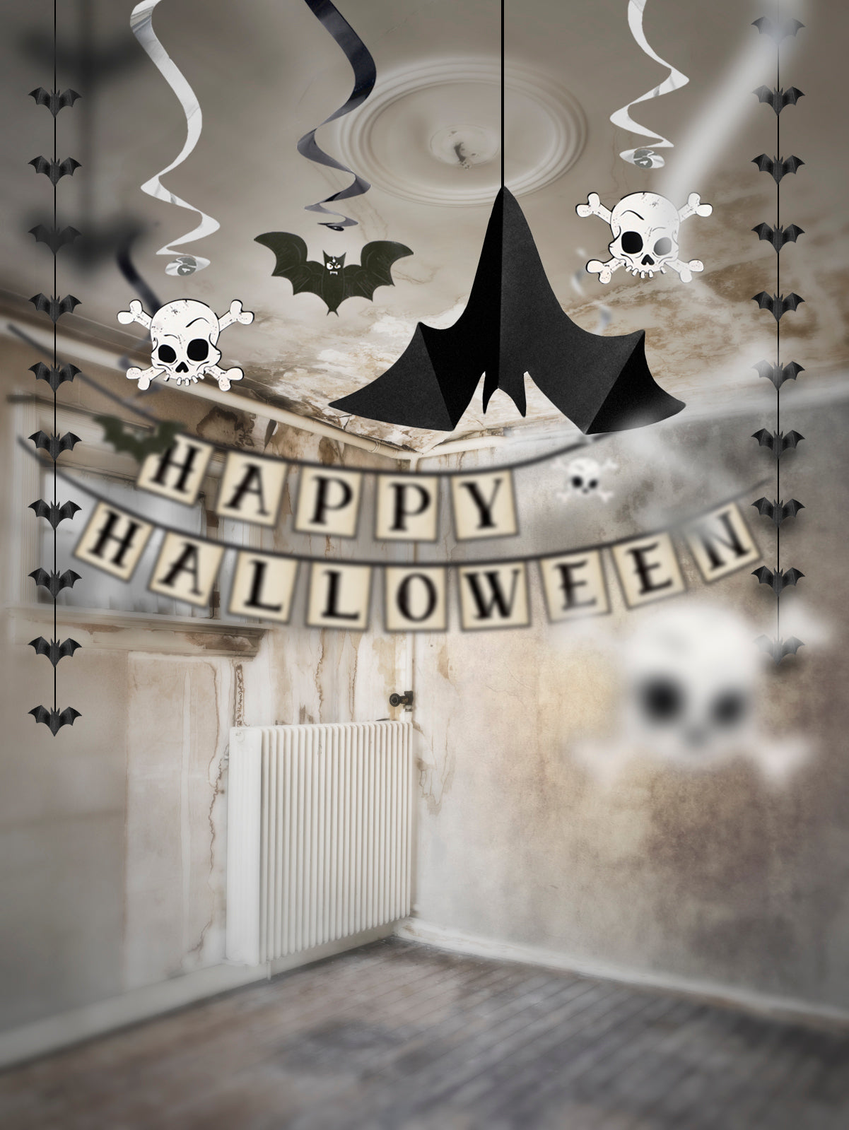Halloween party decoration kit shown hanging in an empty room, featuring a black bat-shaped ceiling ornament, skull and crossbones wall decals, black bat ribbons, a 'HAPPY HALLOWEEN' banner, and white skull streamers.