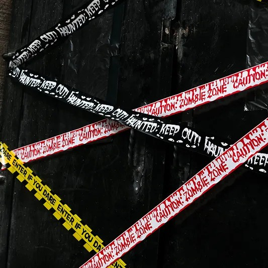 Three diagonal strips of caution tape with 'CAUTION ZOMBIE ZONE' and 'KEEP OUT' text in red, black, and yellow, laid over a dark wooden surface.