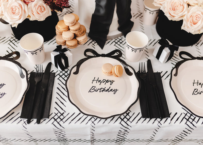 Table setting with 'Happy Birthday' plates, macarons, and flowers on a checkered tablecloth.