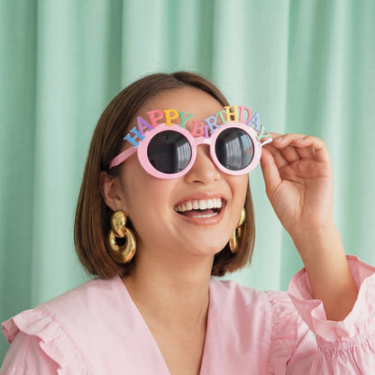 A woman wearing round pink sunglasses with colorful 'HAPPY BIRTHDAY' letter accents on the brow, holding one lens with her hand against a mint green curtain background.