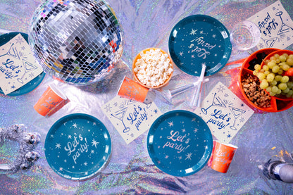 A flat-lay of party supplies on a holographic surface, including blue plates and cups printed with 'Let's party' and starry patterns, orange cups, cocktail napkins, a silver disco ball, popcorn, grapes, and nuts.