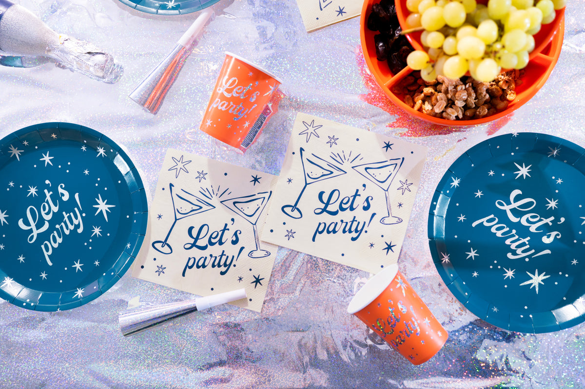 A flat-lay arrangement of party tableware on a sparkly silver surface, including blue paper plates with white script reading 'Let's party!' and orange paper cups with matching text, along with napkins, a party popper, and a bowl of green grapes.