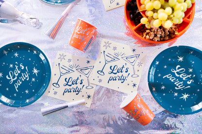 A flat-lay arrangement of party tableware on a sparkly silver surface, including blue paper plates with white script reading 'Let's party!' and orange paper cups with matching text, along with napkins, a party popper, and a bowl of green grapes.