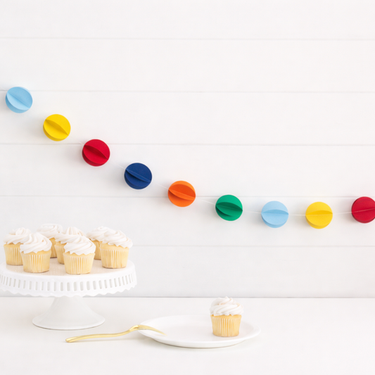 Rainbow coloured 3D circle garland hanging above a cupcake table