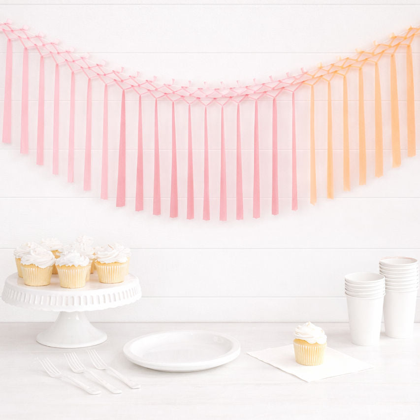 Pink and peach tissue tassel garland hanging above a party table with cupcakes and white cake stand