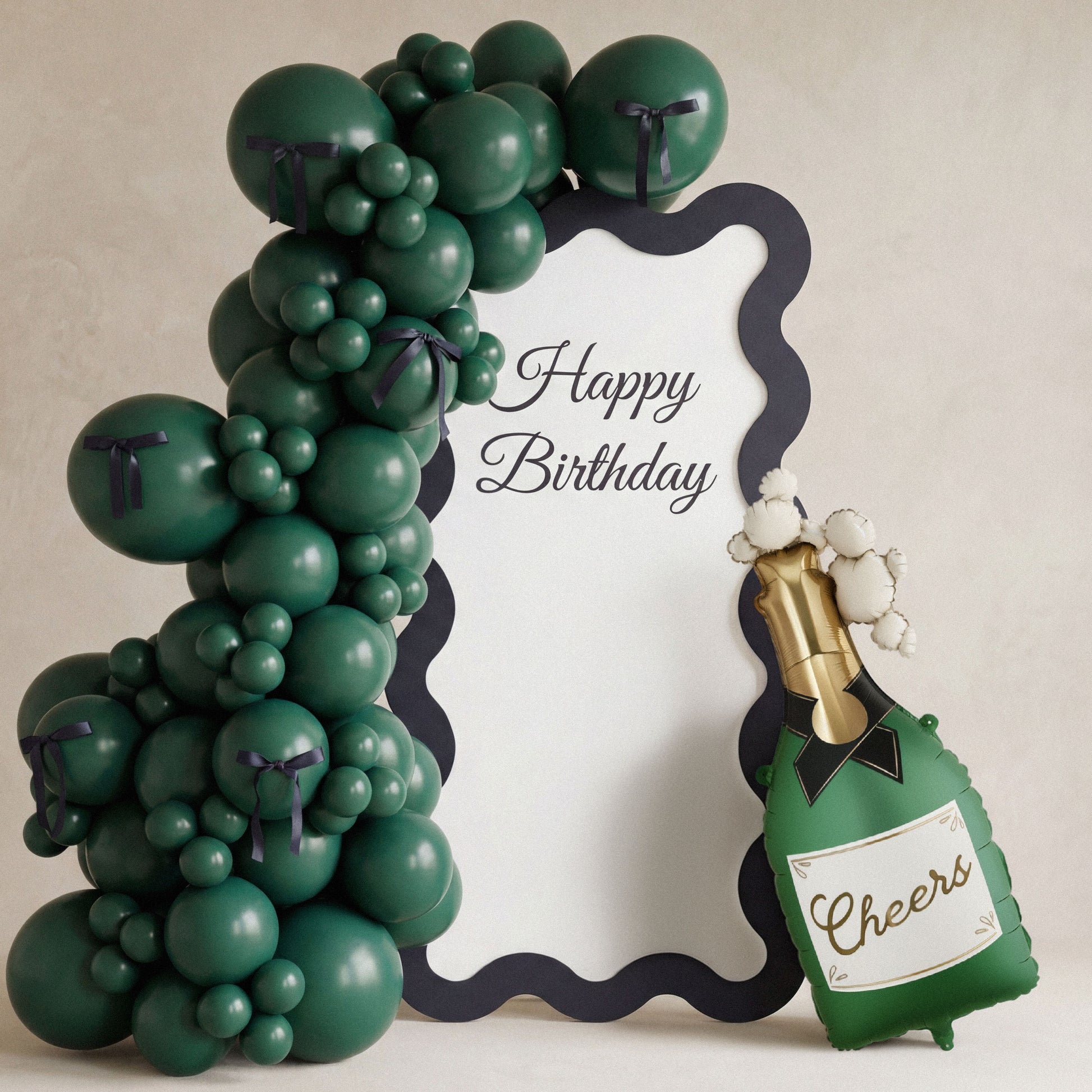 Green balloon arch with a 'Happy Birthday' sign and a bottle-shaped balloon with 'Cheers' text on a beige background.